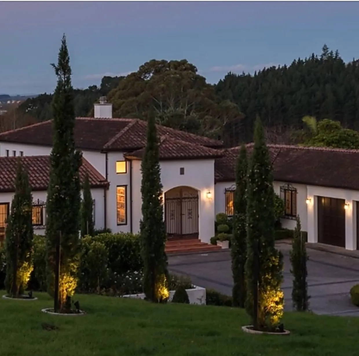 A Spanish-style Auckland home at dusk, with clipped hedging, uplit cypresses and lawn running down to the drive